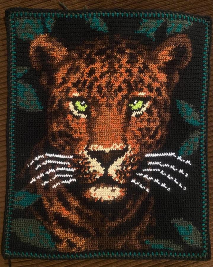 Brown and black jaguar crochet wall hanging ideas with green leaves from @crochetedbylarissa. Detailed animal portrait on dark background.