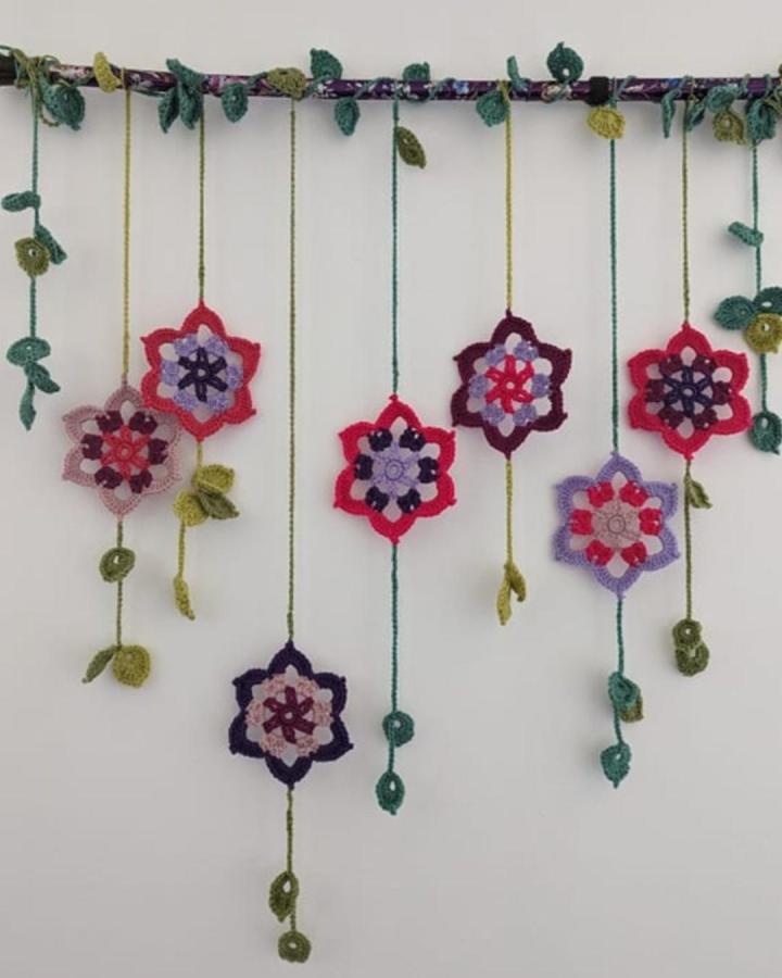 Colorful crochet floral garland with leaf accents hanging from fabric-wrapped branch from @opheliatalks. Handmade crochet wall hanging ideas with nature-inspired details.