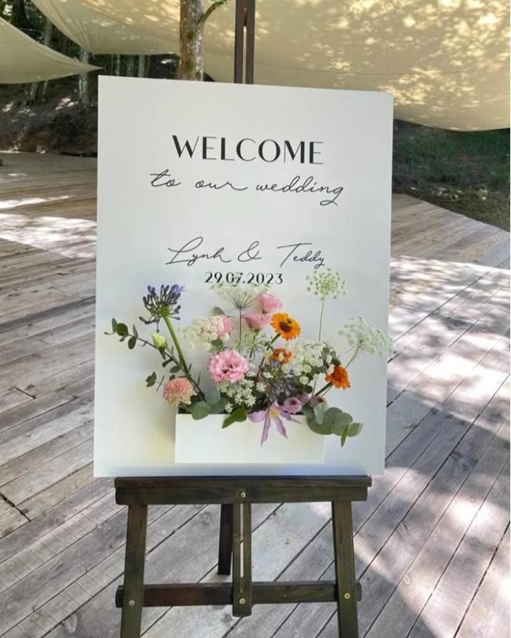 White board wedding sign with colorful mixed flowers in a box from My Valentine Creative Studio bright and welcoming diy wedding welcome sign ideas on wooden easel outdoors