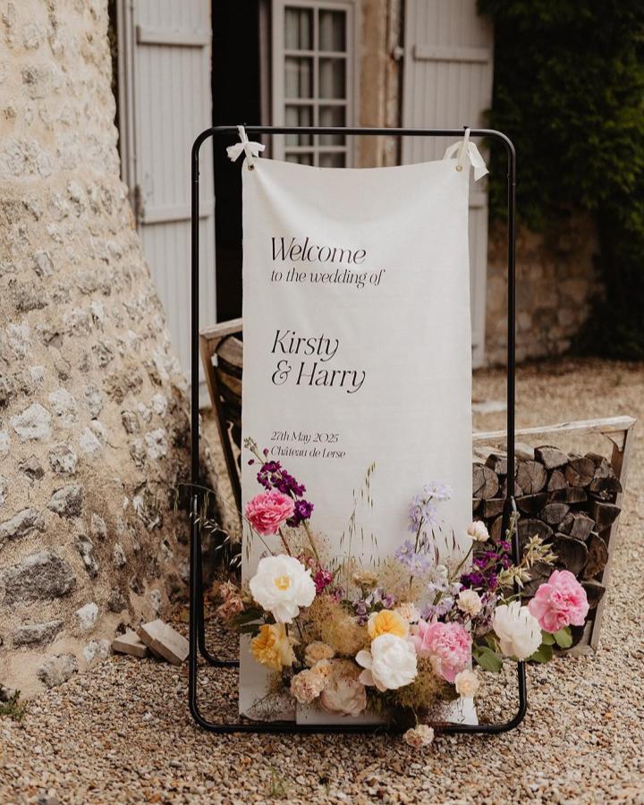 Cream fabric banner on black metal frame with fresh pink and purple flowers from @bewilderly. Rustic diy wedding welcome sign ideas.