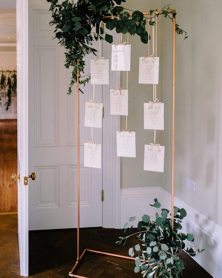 Copper frame with hanging cards and greenery from Boho Weddings showcasing diy wedding welcome sign ideas with natural eucalyptus accents and ribbons