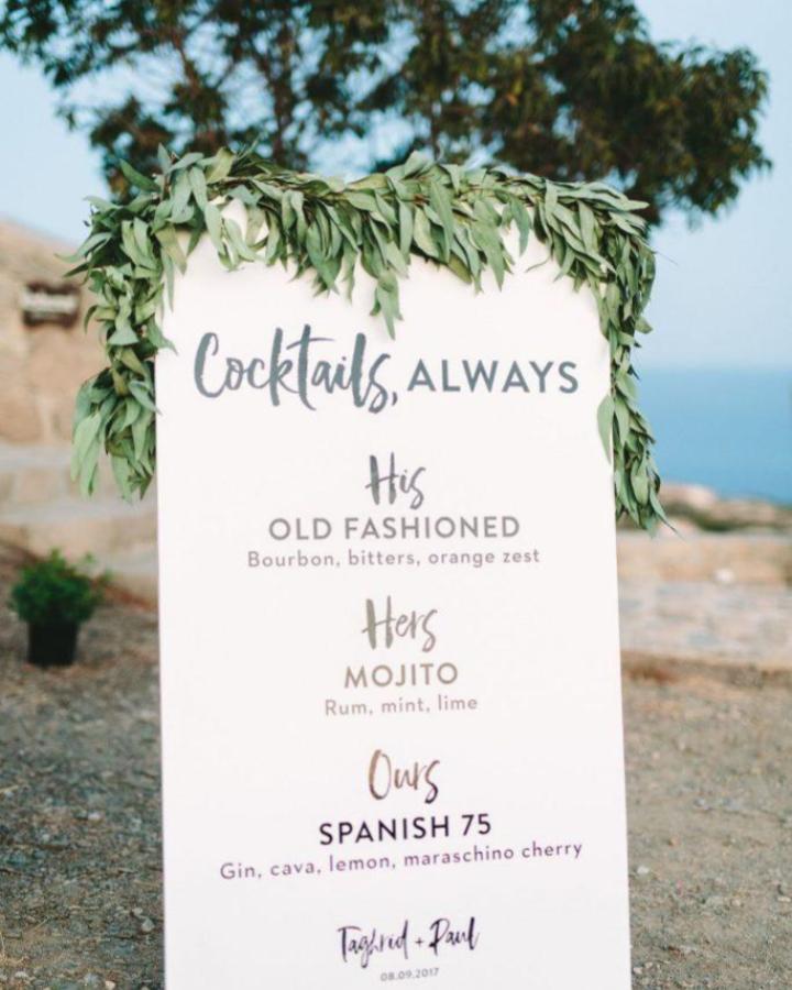 White cocktail menu sign with green leaf garland on top from Style Me Pretty. Outdoor rustic wedding welcome sign with drink options listed.