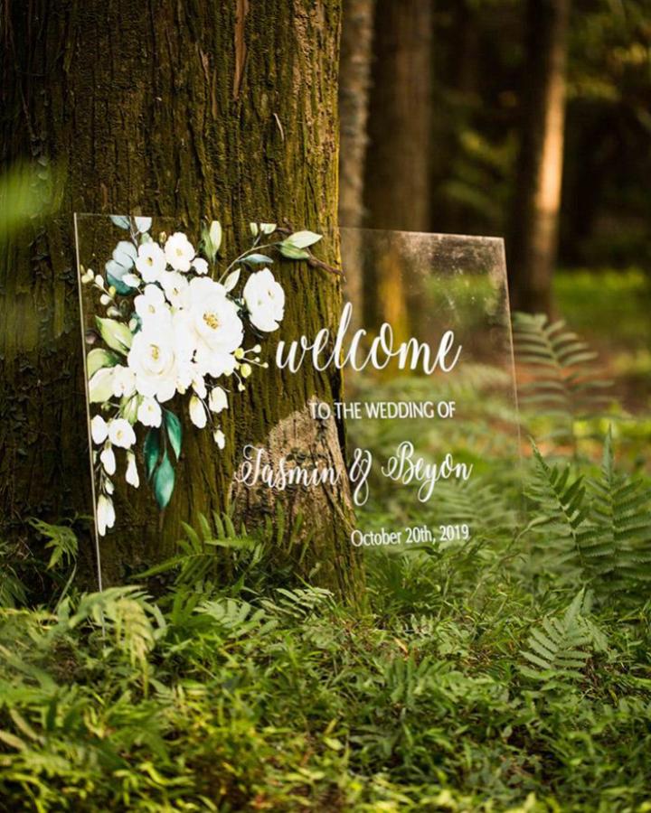 clear acrylic wedding welcome sign with white floral paint in forest setting from Elegant