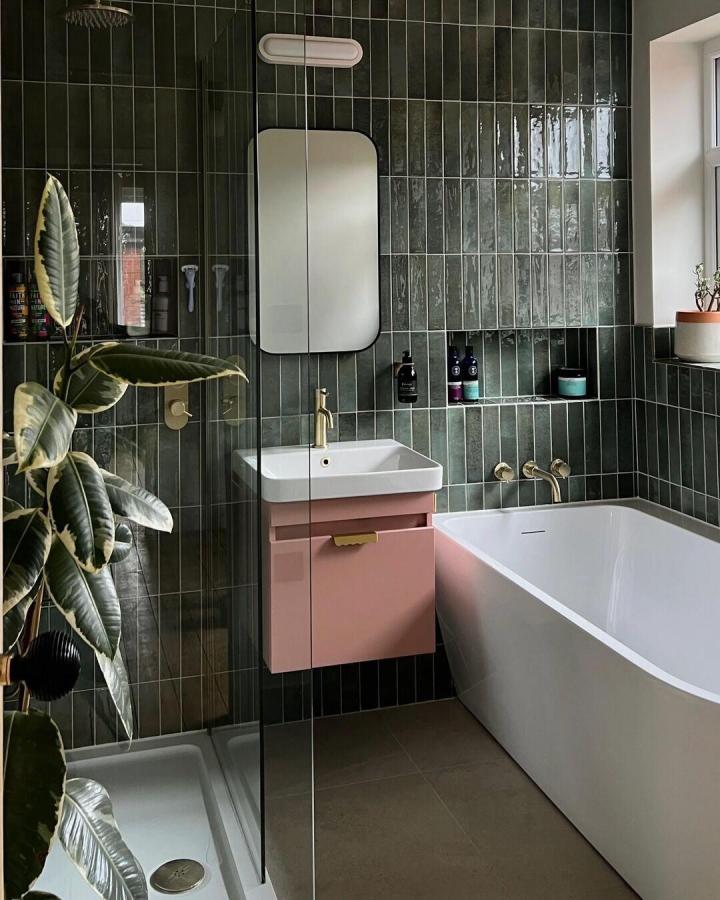 Glossy forest green tiled bathroom with pink vanity and brass fittings from @firedearthuk showing green bathroom decor ideas.