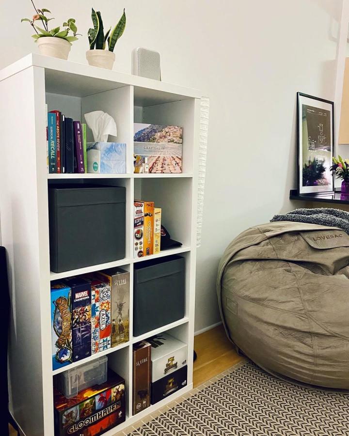 White Ikea Kallax bookshelf with gray bins and colorful board games next to a beige beanbag from @boardcertifiedgames styled for ikea kallax bookshelf styling ideas.