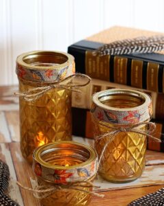 Golden quilted glass mason jar candle holders with burlap leaf ribbon and twine bows from House of Hawthornes glowing warmly on rustic wood surface.