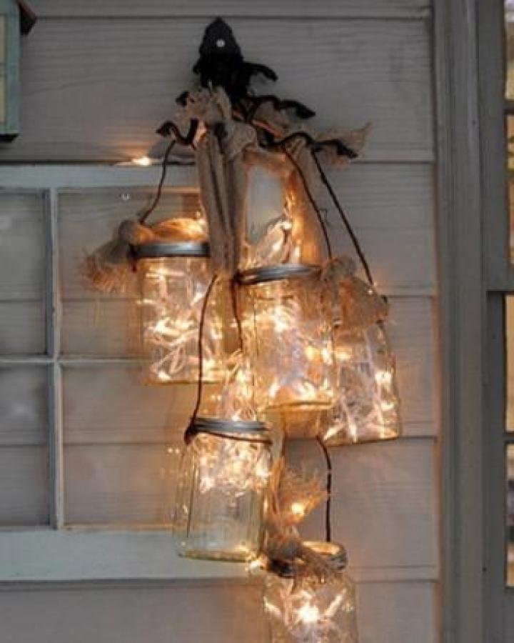 Clear glass mason jar lights hanging with burlap ribbons creating rustic ambient lighting for adults craft from All Things Heart and Home. Clustered glowing jars with soft fairy lights inside.