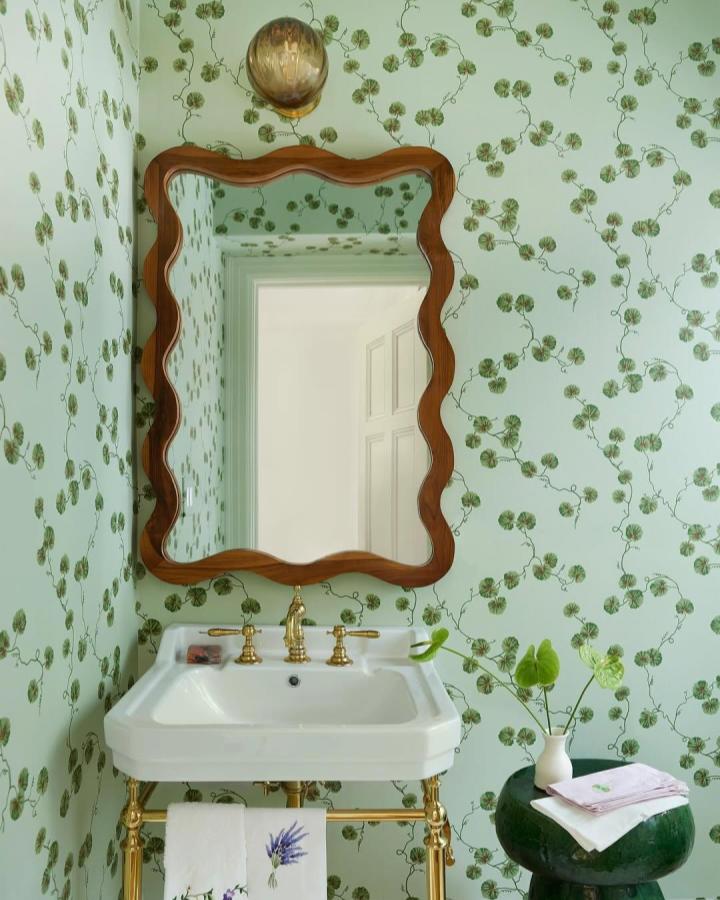 Soft green botanical wallpaper with delicate vines and clovers in a small powder room from @colleensimondsdesign featuring brass fixtures and a scalloped mirror frame.