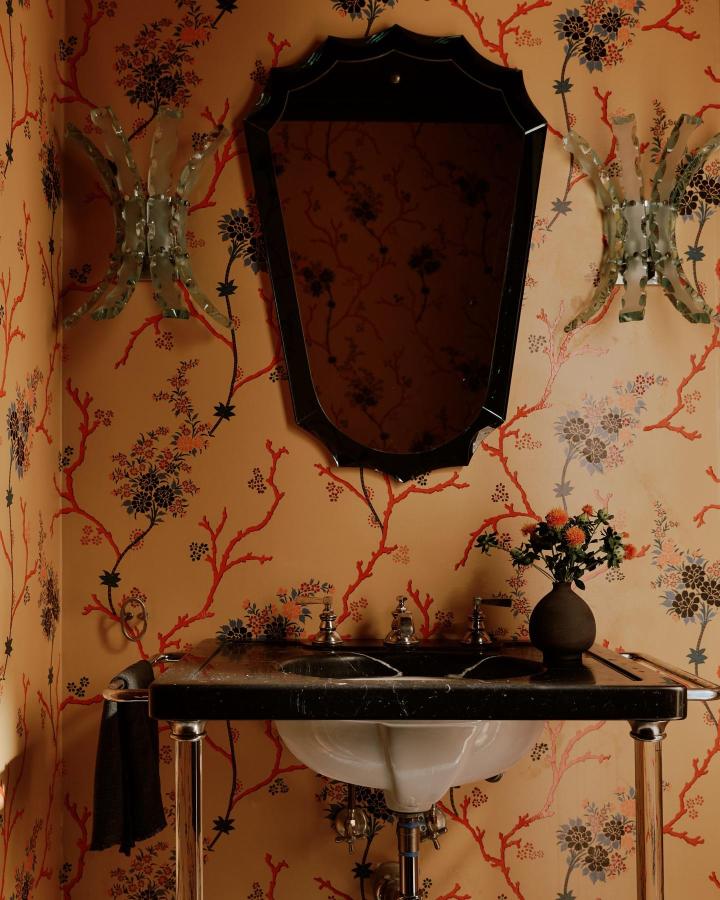 Apricot botanical wallpaper with coral branches and dark floral accents in a small powder room by @chusedandco. Elegant vintage style with marble sink and sculptural sconces.