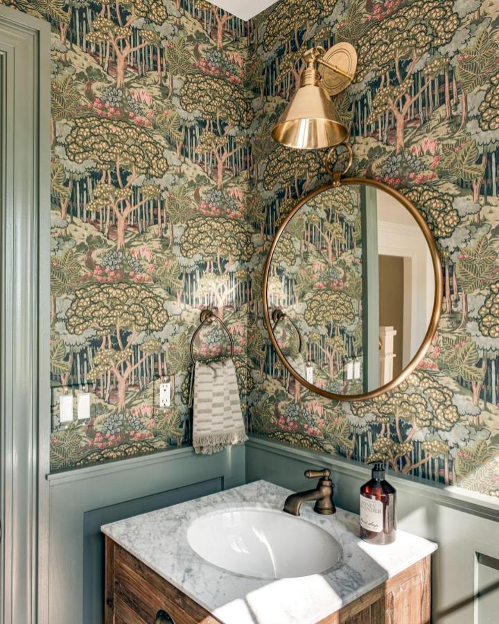 Vintage forest patterned wallpaper in a small powder room featuring brass mirror and green wainscoting from @wainteriordesign creating cozy woodland vibe.