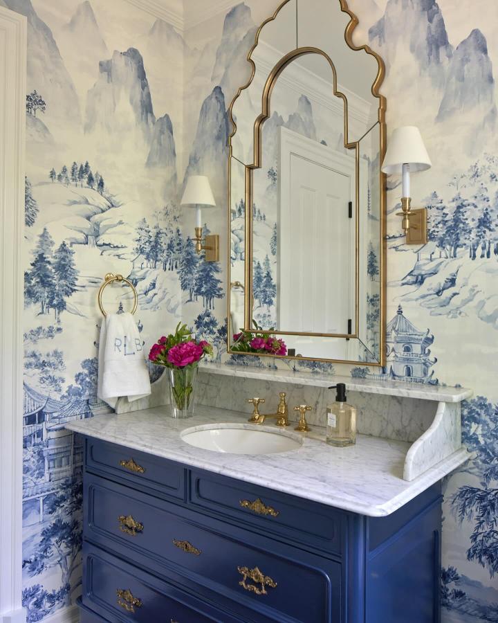 Blue and white scenic wallpaper with navy vanity and gold accents in a small powder room from @amalkapeninteriors. Elegant and cozy design.