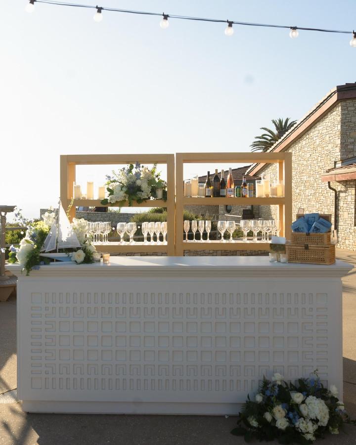 White minimalist bar with wood shelves and floral accents from @champagnedreams.co wedding bar ideas with outdoor setting and string lights