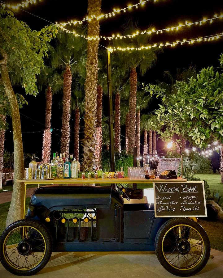 Vintage navy blue car bar cart under string lights from @peppelasala. Wedding bar ideas with palm tree background and cozy outdoor vibe.