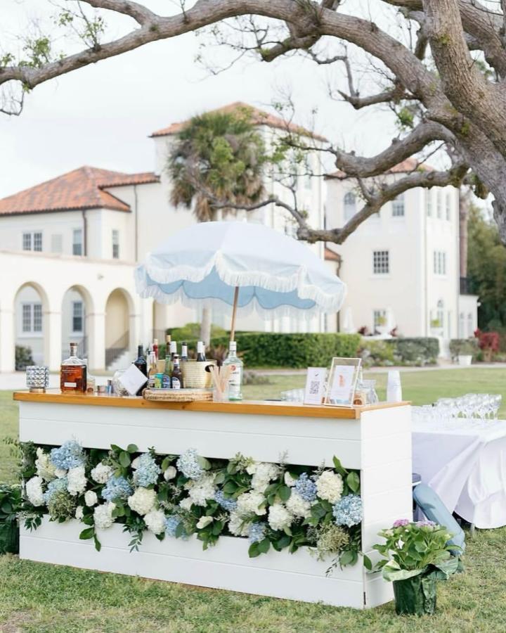 White wooden outdoor bar decorated with blue and white hydrangeas and greenery from @the.wandering.whale for wedding bar ideas outdoors. Elegant floral design with umbrella.