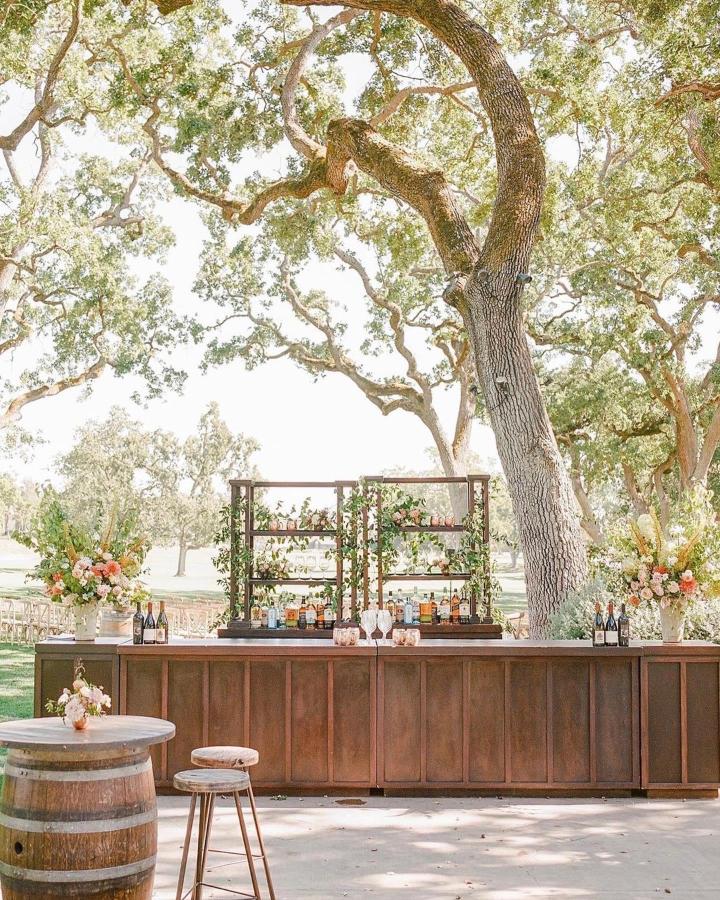 Rustic wooden wedding bar ideas from @encoreeventsrentals with greenery and floral accents under large oak trees outdoors.