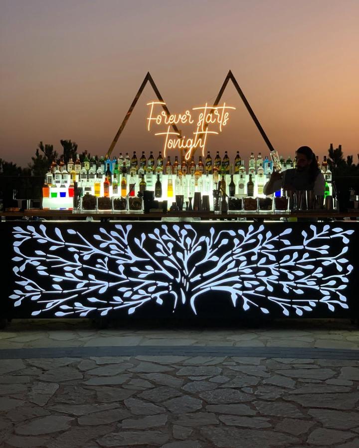 Glowing white branch pattern on dark wooden wedding bar from @bottlefly.lb with neon sign and bottles at twilight wedding bar ideas setup.