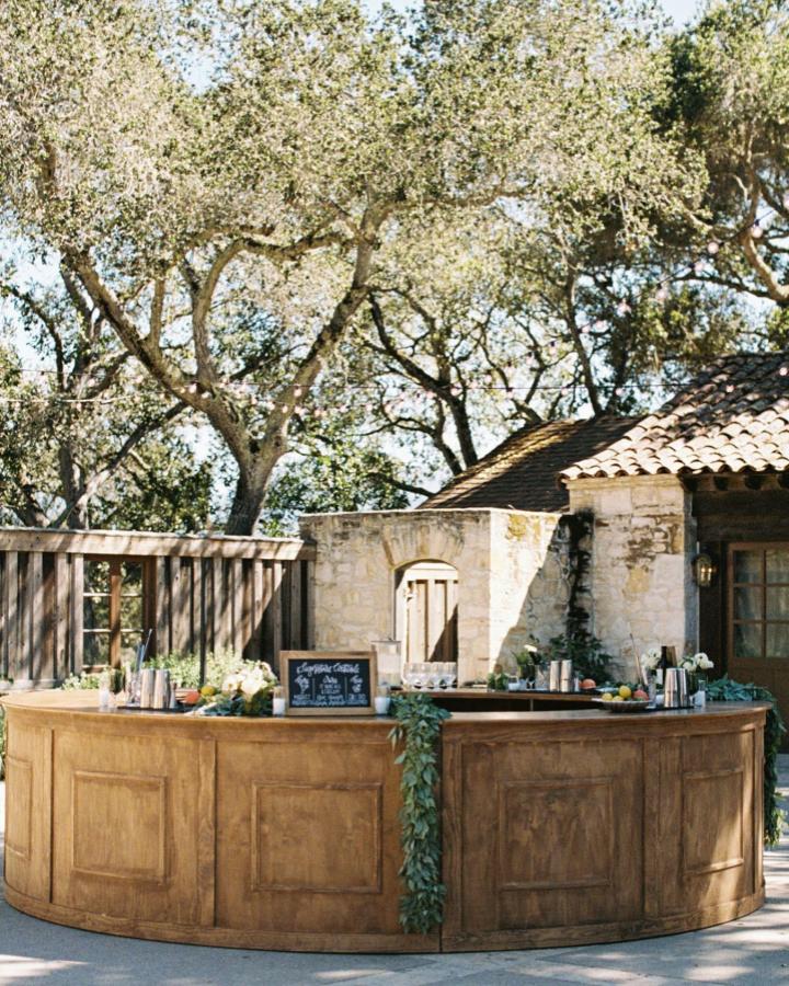 Rustic wooden half moon bar with greenery and chalkboard from @standardeventrentals for wedding bar ideas outdoor setup surrounded by trees