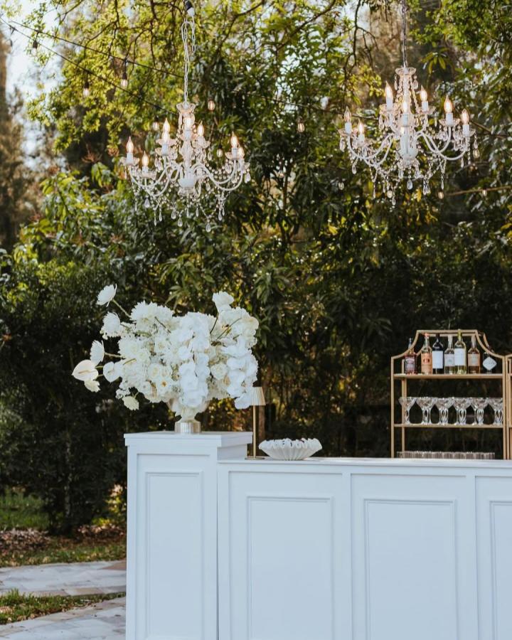 White crystal chandeliers hanging over a classic white paneled bar with gold wine rack and white flowers from @boozeandbubbless wedding bar ideas outdoors.