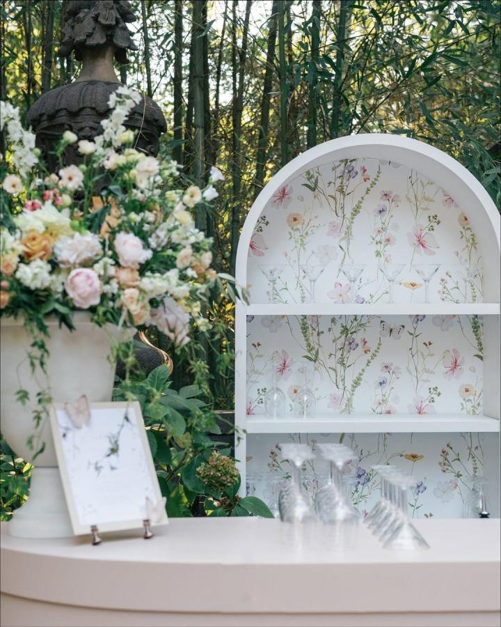 Soft pastel floral wallpapered curved shelves and delicate glassware from @party.bound. Elegant outdoor wedding bar ideas with vintage touches.
