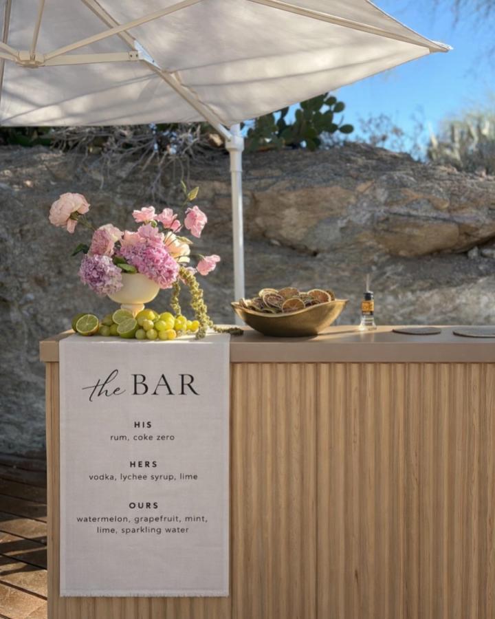 Natural linen menu banner draped over wooden bar with pink flowers and fruit from @benjimobilebar for wedding bar ideas outdoor setup.