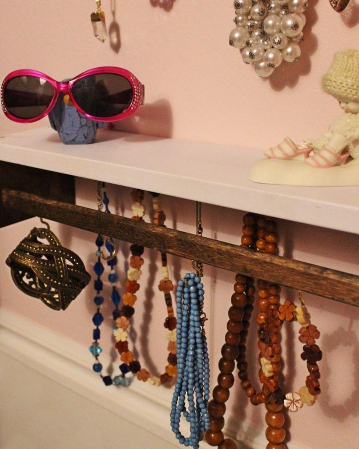 Pink wall with wooden bar necklace rack and colorful beaded necklaces hanging below from Homedit. Rustic jewelry organizer made from a repurposed spice rack.