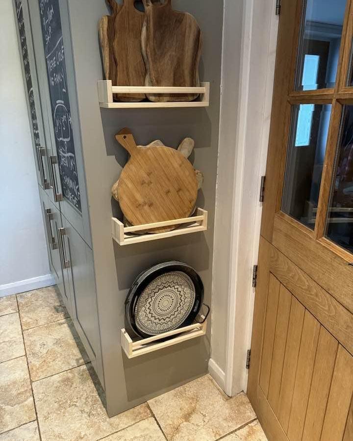 Wooden wall-mounted shelves holding cutting boards and trays in a kitchen corner by Clare Wallace practical kitchen storage solution.