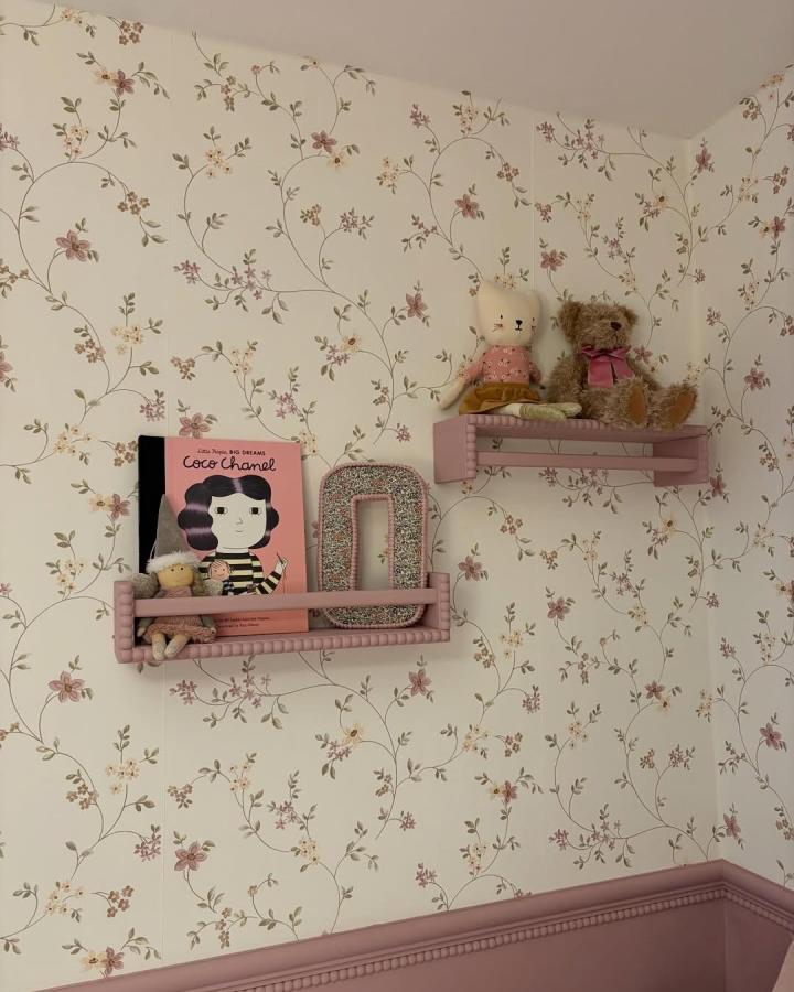 Soft pink painted spice rack shelves mounted with toys and books in a floral wallpapered room from @the.dobsonhome cozy display hack idea