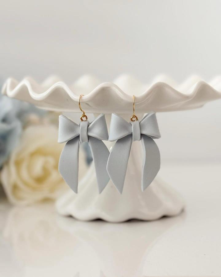 Soft gray bow-shaped earrings with gold hooks on a white ceramic stand from @wildhoneyclay. Elegant polymer clay accessory idea.