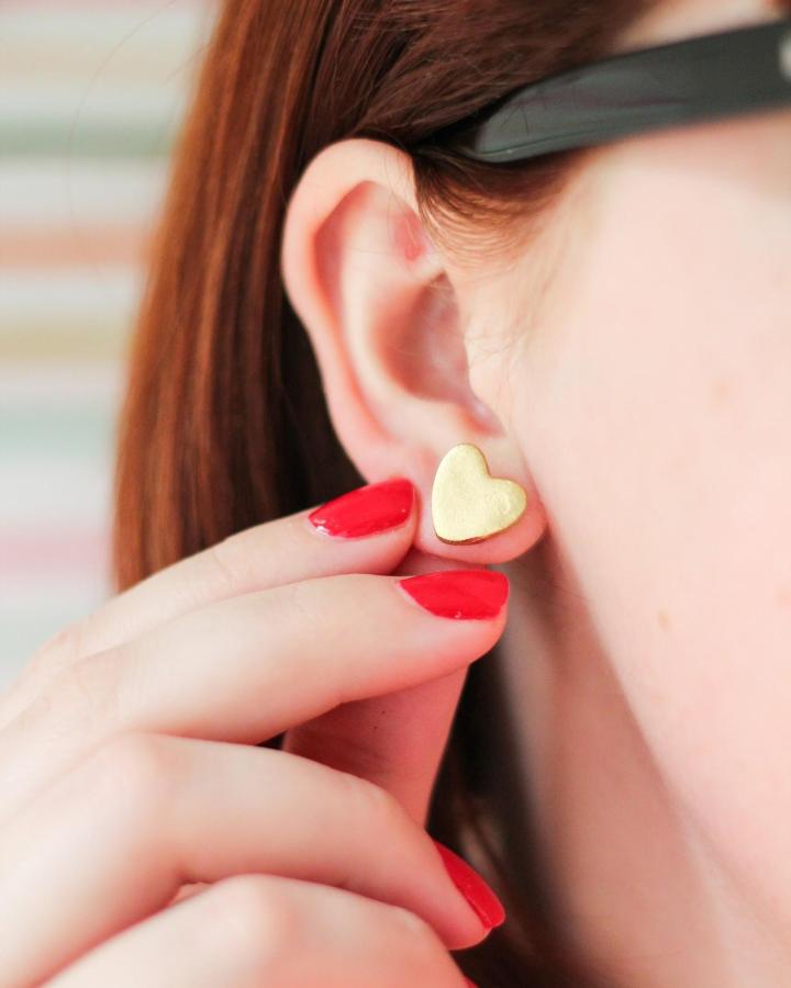 Gold heart-shaped polymer clay stud earrings held near ear with red nail polish polymer clay earrings ideas from the crafted life.