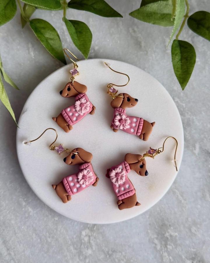 Brown dachshund earrings with pink sweaters and floral details crafted from polymer clay by @sarah.cotta.clays resting on a white dish.