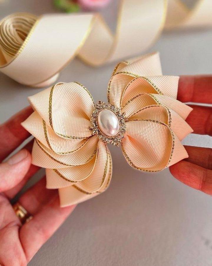 Cream ribbon bow with gold trim and pearl centerpiece held in hands by @madebydianasahafe. Elegant handmade accessory with vintage charm for crafts.