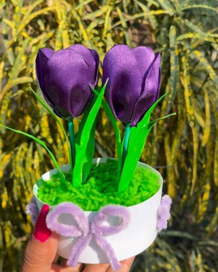 Purple ribbon tulips with green leaves in a white pot decorated with lilac pipe cleaner bows held outdoors by hand from @creative_cornerbyme. Bright and textured floral ribbon craft for adults.