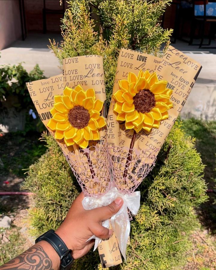 Yellow sunflower ribbon bouquet wrapped in love-print kraft paper held outdoors by hand. Bright and textured floral craft with brown center and white bow.