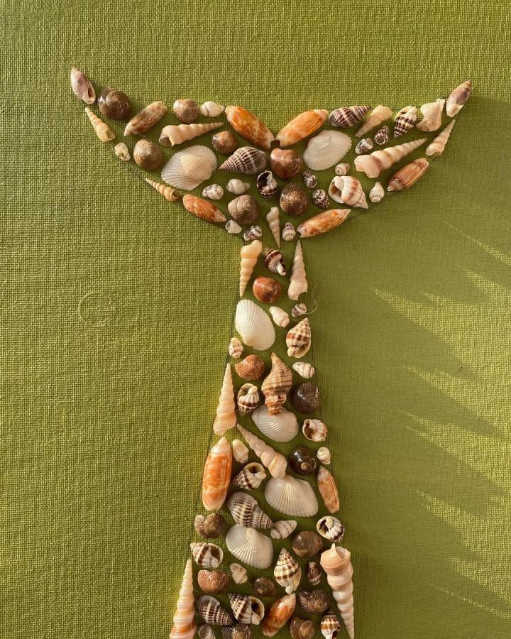Olive green textured background with varied brown and cream seashells arranged in a whale tail shape by @artisanshonali for coastal inspired crafts.
