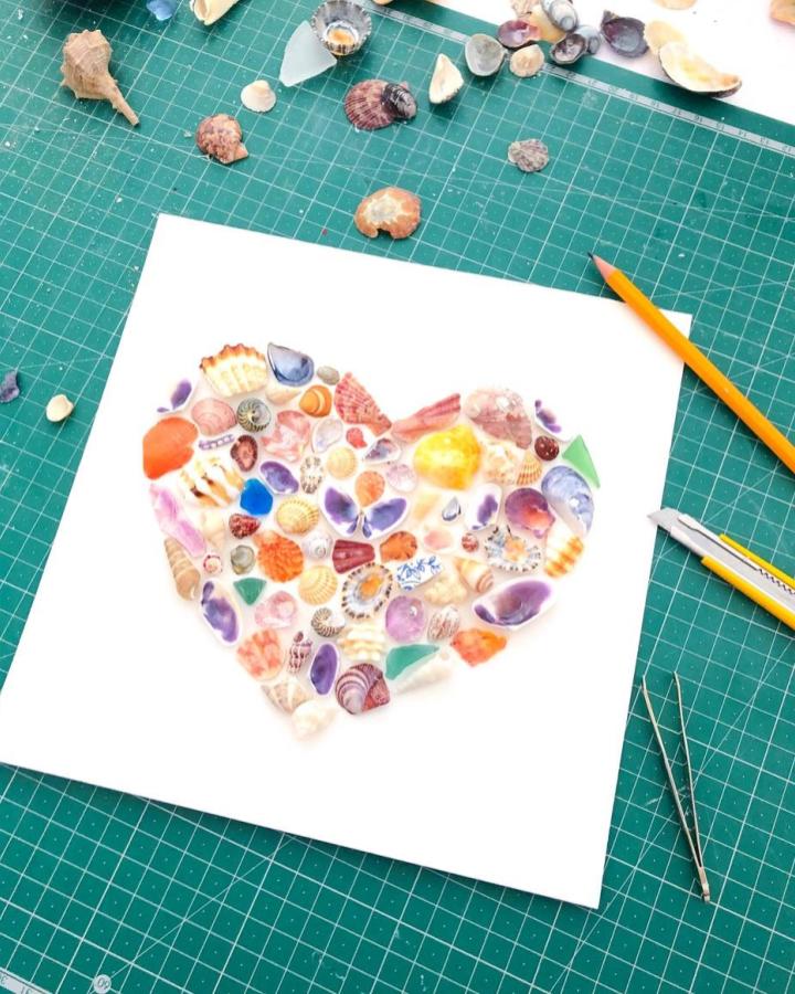 Colorful seashell and sea glass heart collage with crafting tools on green cutting mat by @seabylou. Handmade coastal-inspired craft art.
