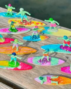 Miniature colorful surfer figurines on glittery boards with bright summer hues by @craftalux, perfect for sunny craft displays.
