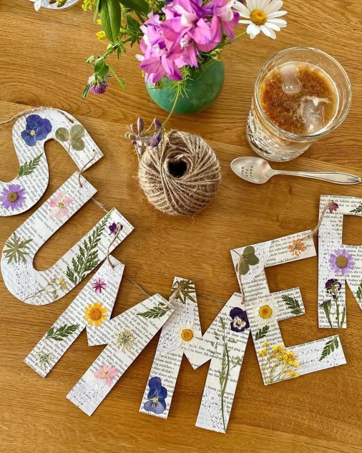 Pressed flower decorated paper banner spelling summer with twine and lace on wooden table by @lindamydlak. Rustic natural craft idea with vintage flair for warm seasons.
