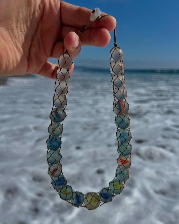 Sea glass marbles necklace handcrafted with mesh netting hanging against ocean waves perfect for seaside craft inspiration by @seaglasshoe