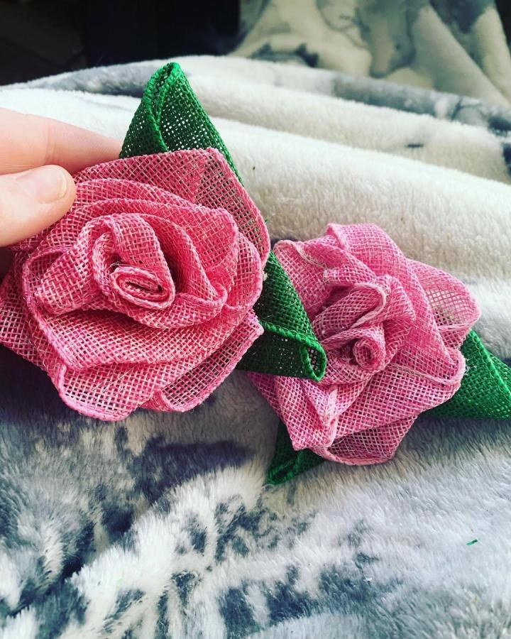 Pink and green burlap handmade roses with textured petals and leaves from @amandasflairart displayed on fluffy fabric background.