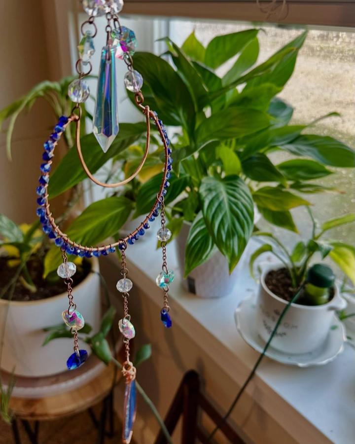 Copper wire hoop with blue and iridescent beads hanging near green plants from @kmcjewelrydesign. Bright, intricate handcrafted suncatcher with natural light effects.