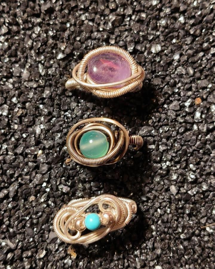Polished gemstone rings with copper wire wrapping from @malikasrealm featuring amethyst, turquoise, and green stones on textured background.