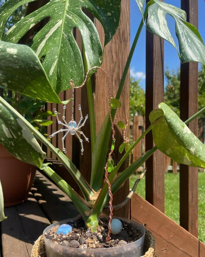Copper wire sun catcher plant stake with sparkling beads from @lunastarastudio hanging on leafy green potted plant outdoors in sunlight.