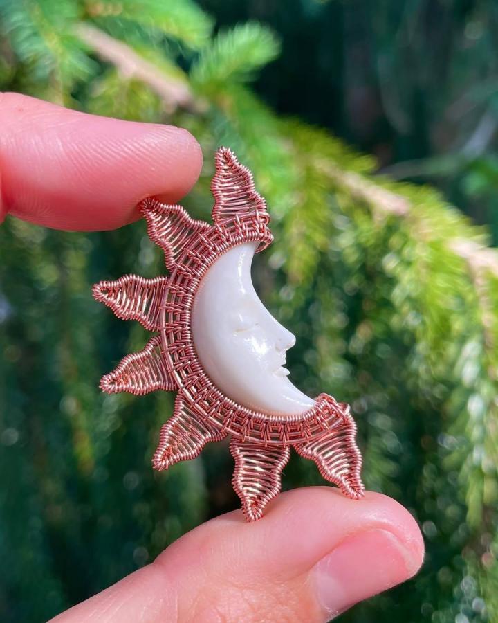 Copper wire sun and moon pendant featuring white crescent stone held between fingers by @wickedwirecrafts. Intricate wire weaving details.