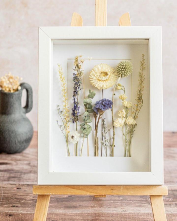 Cream and lavender pressed wildflowers arranged in a white minimalist frame by @magentabloomsnz elegantly displayed on a wooden easel.