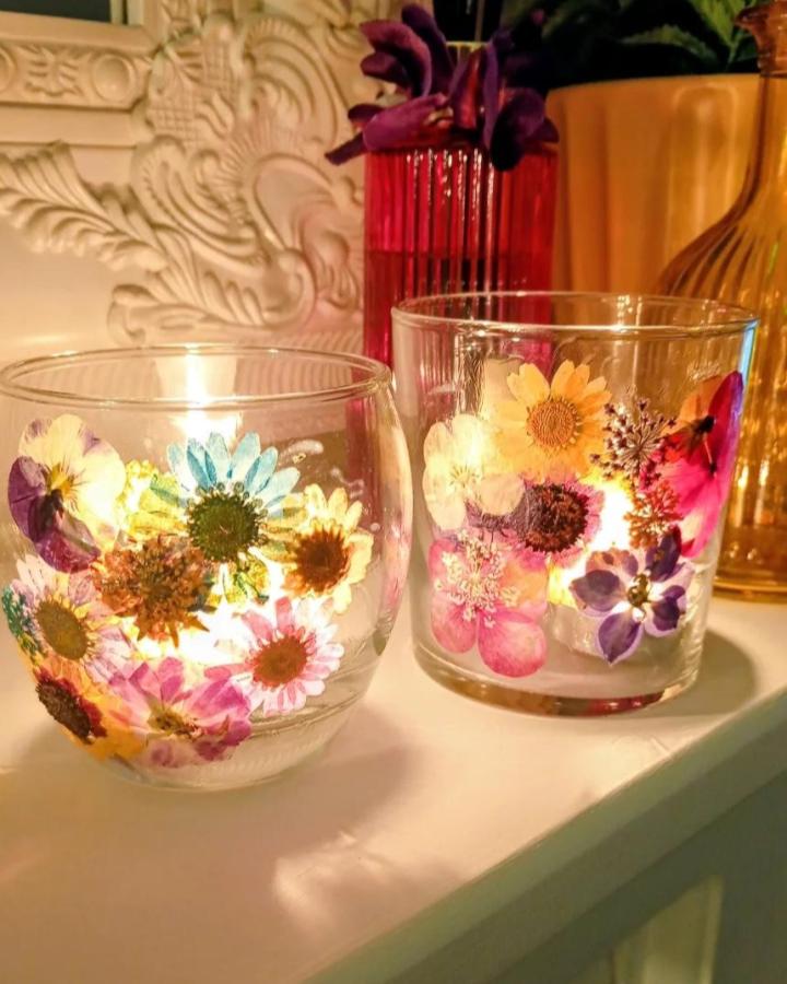 Pressed colorful flower decorated glass candle holders glowing warmly on a white surface from @victoriasglasses. Dried flower crafts creating cozy light and texture.