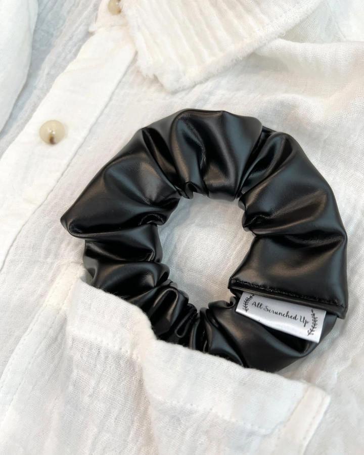 Black shiny faux leather scrunchie crafted by @allscrunchedup.pp placed on white linen shirt pocket. Glossy and stylish hair accessory.
