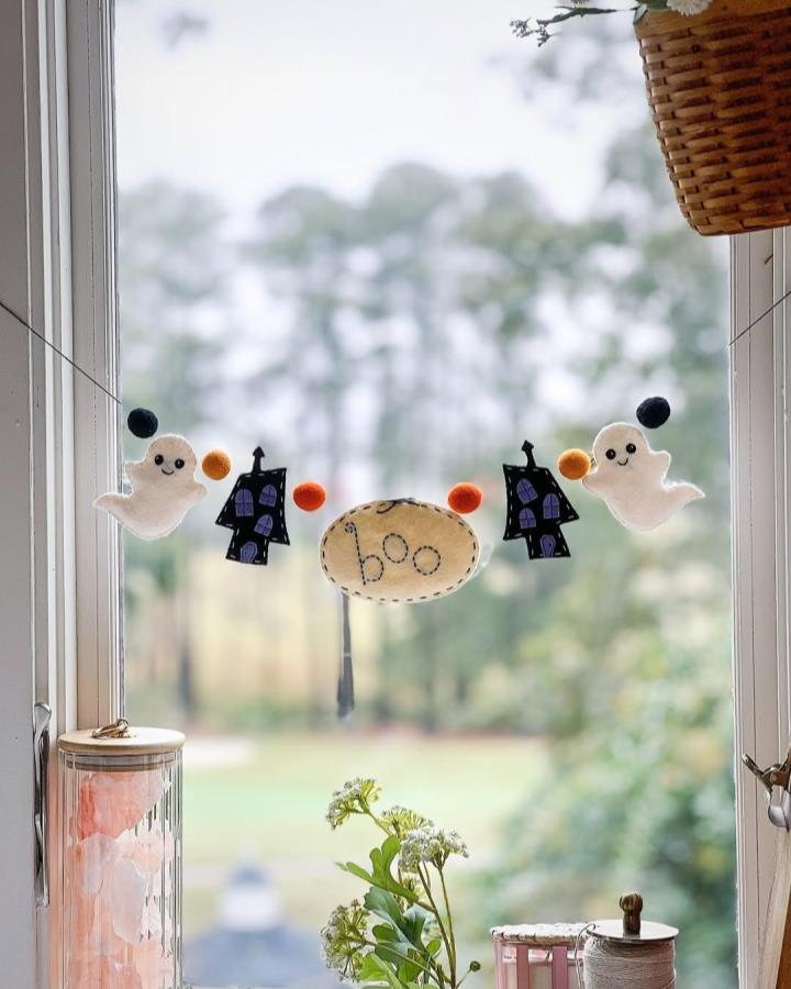 White and purple felt Halloween garland with ghosts and houses by @amysinibaldi hanging in window for seasonal craft decor.