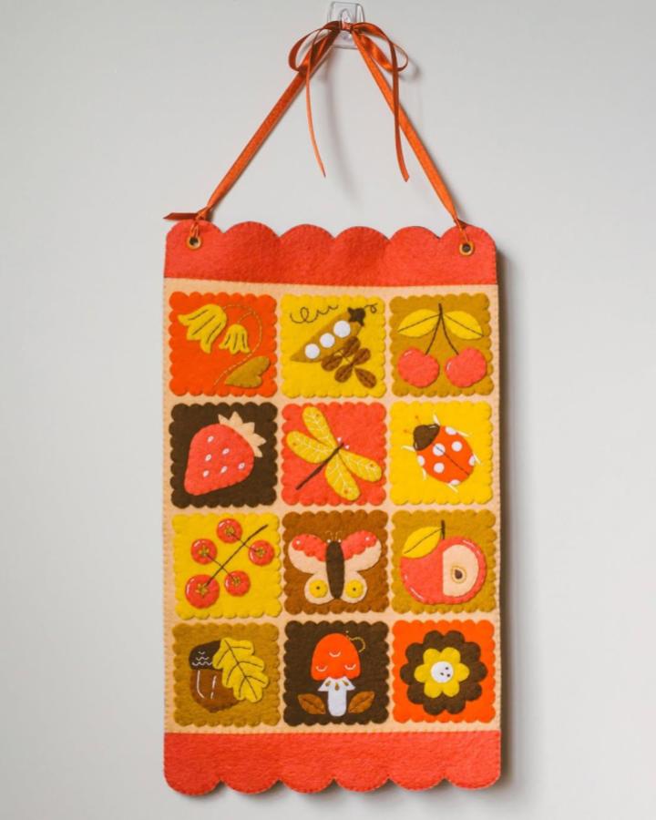 Orange and yellow felt patchwork wall hanging from @ferntales_toys with cute applique designs of fruit and insects, cozy handmade decor.