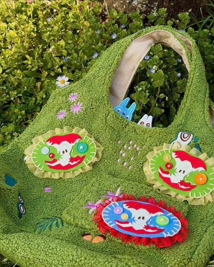 Green textured bag with colorful ruffled felt pins by @beeb0z. Playful stitched and button details add whimsical charm.