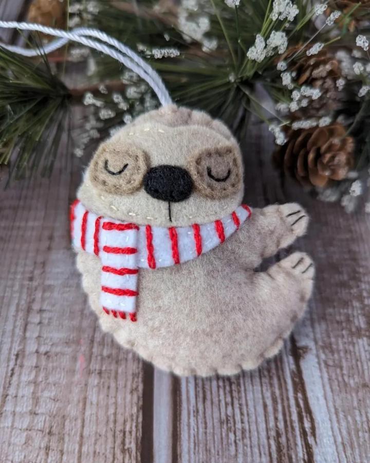 Beige felt sloth ornament with red striped scarf on rustic wood, creative handmade holiday craft from @olivethisfelt. Cozy and cute design.